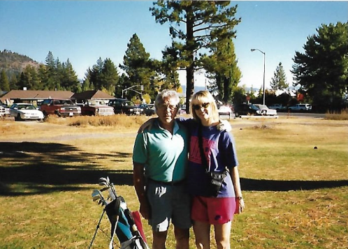 In Loving Memory of Dad: This photo captures a heartwarming moment between a father and his daughter, standing arm-in-arm outdoors on a sunny day. The scene is peaceful, with a backdrop of tall trees, mountains, and a golf course. The father's gentle presence and the daughter's affectionate smile reflect the deep bond they shared, a cherished memory of love and togetherness.